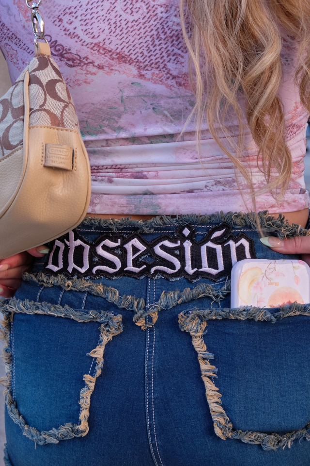 A person with long, wavy blonde hair wears a distressed denim skirt with "obsesión" embroidered above the back pockets, carrying a beige designer handbag.